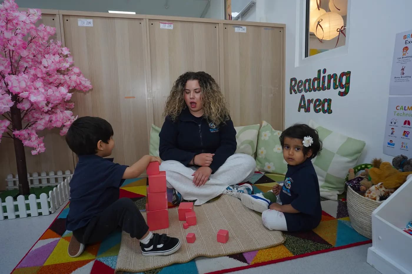 Teacher with two children building with red Montessori cubes by the cherry-blossom reading area at Blue Nest Borehamwood